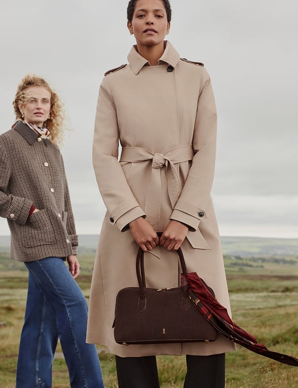 Woman in a beige trench coat with brown belt holding a handbag, standing outdoors with rolling hills in background.