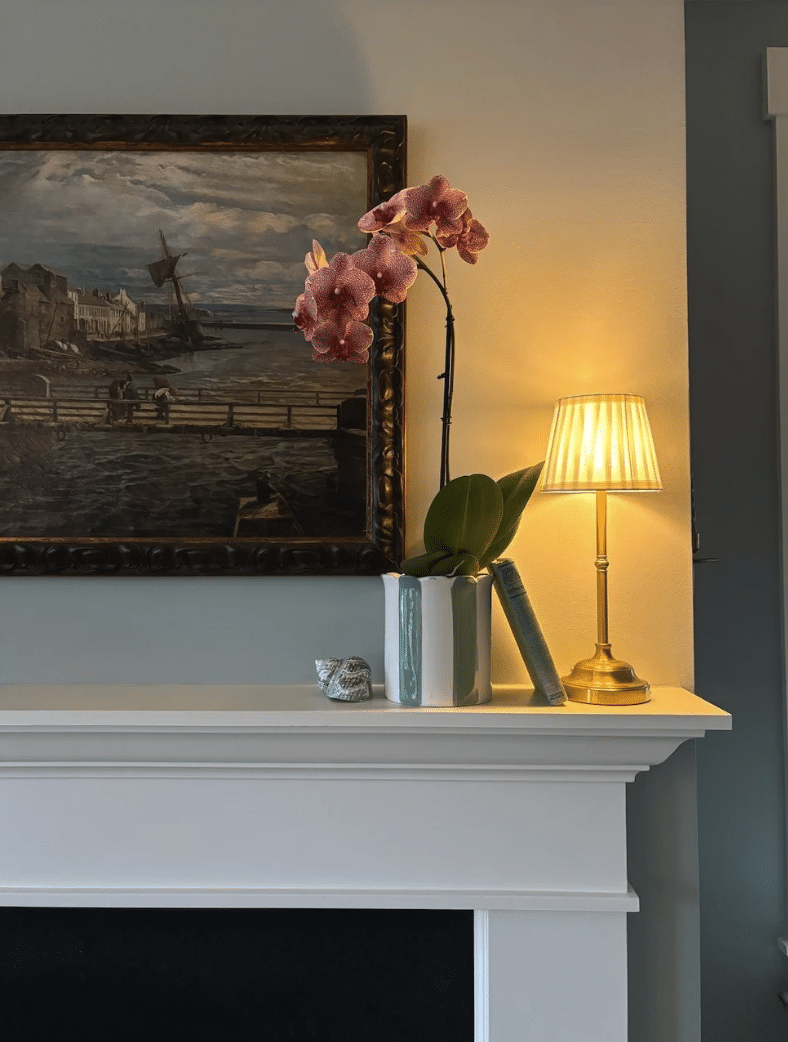 Fireplace mantel decorated with pink orchid in vase, stacked books, brass table lamp, and framed coastal painting