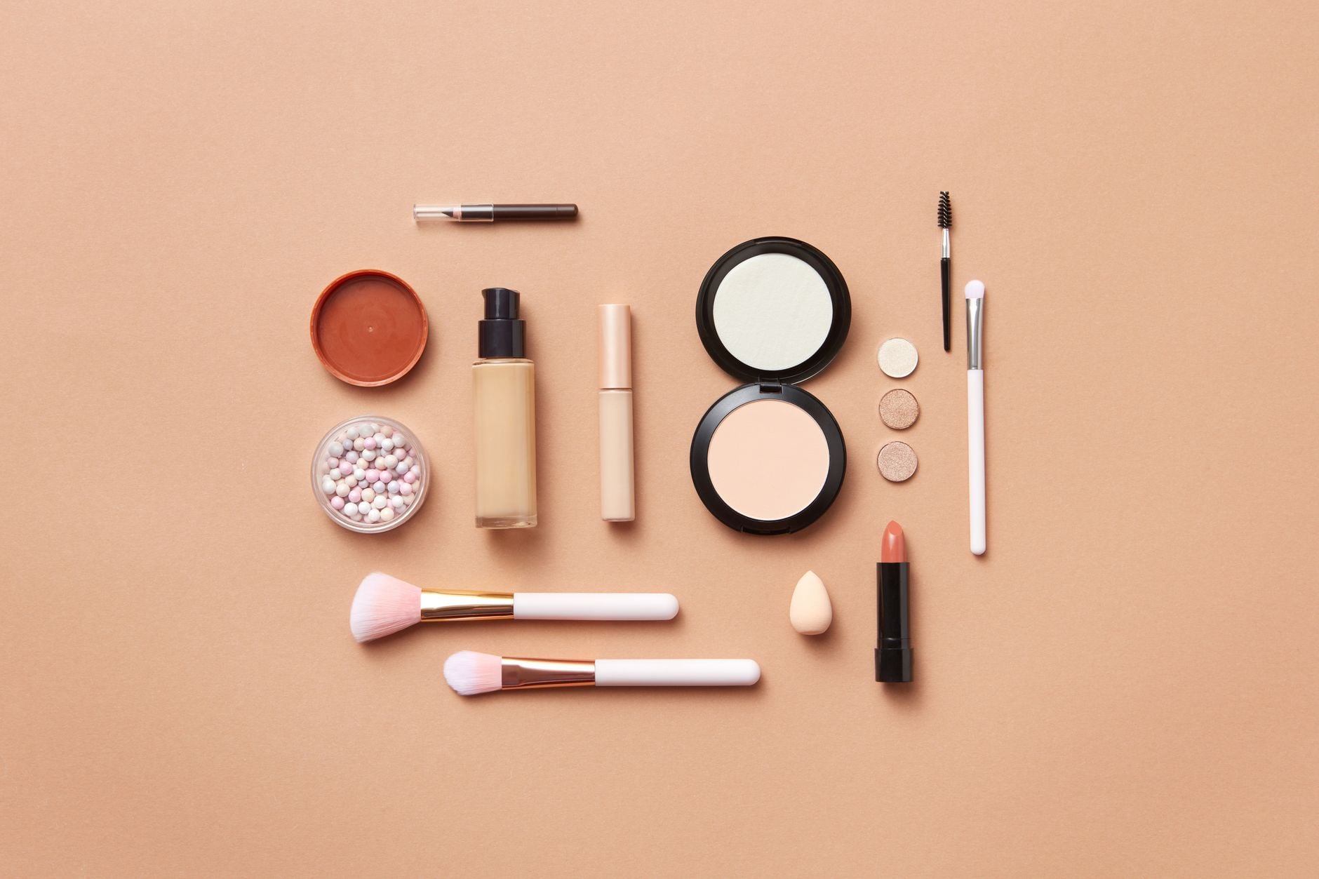 Flat lay of makeup products including foundation, powder, brushes, lipstick, and eyeshadow on beige background