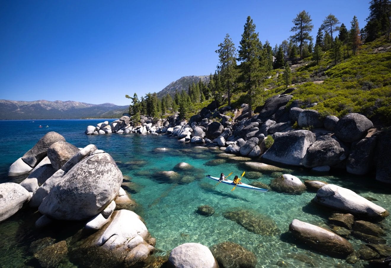 Paddleboarders on clear blue lake with granite boulders, conifer forest, and mountains in background