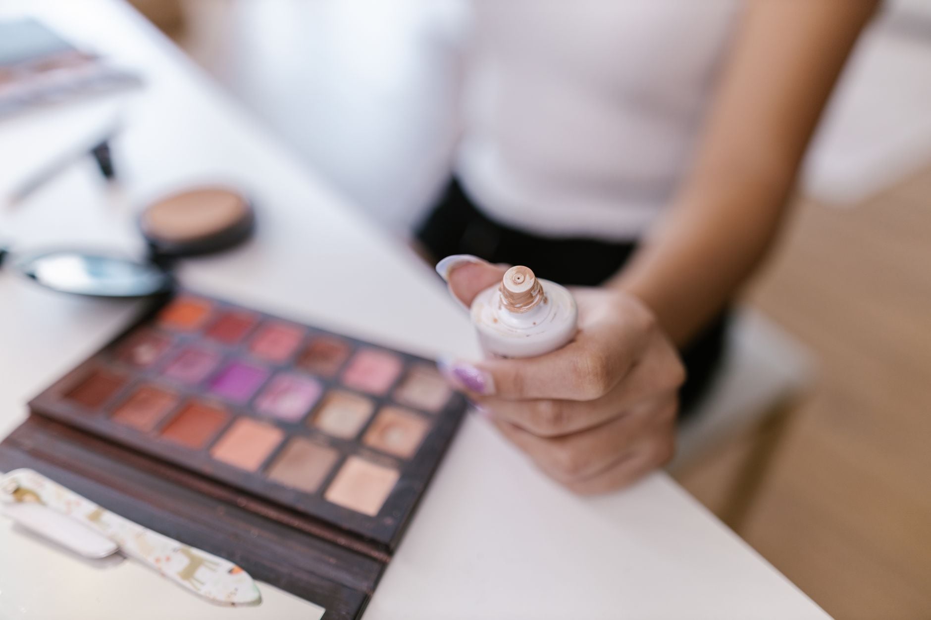 Person holding open foundation bottle with makeup palette and brushes on table