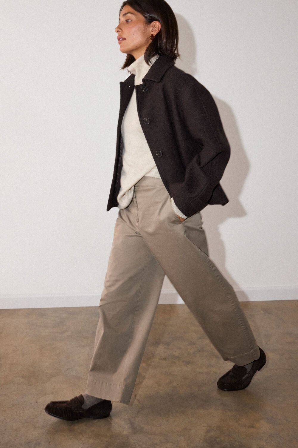 Woman with shoulder-length dark hair wearing black blazer over cream turtleneck and beige trousers, walking