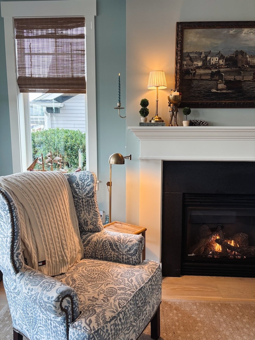 Upholstered wingback chair with blue damask pattern near fireplace and window with brown woven blinds