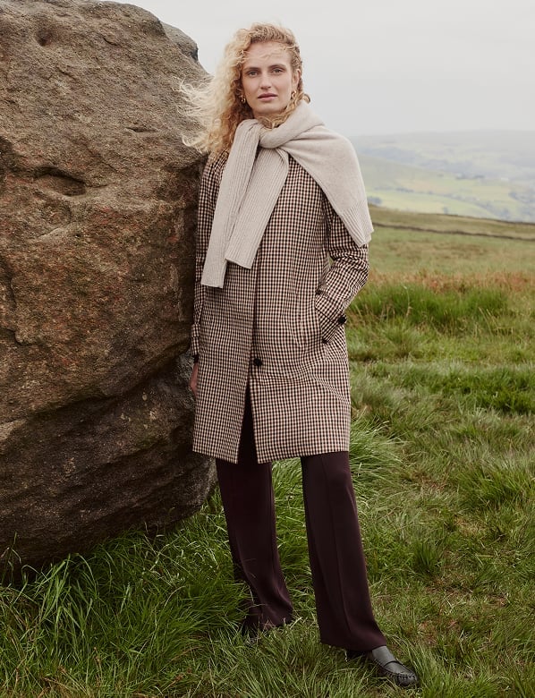 Woman with blonde curly hair wearing a brown gingham coat and cream scarf, standing by a large rock in a rural hillside landscape.