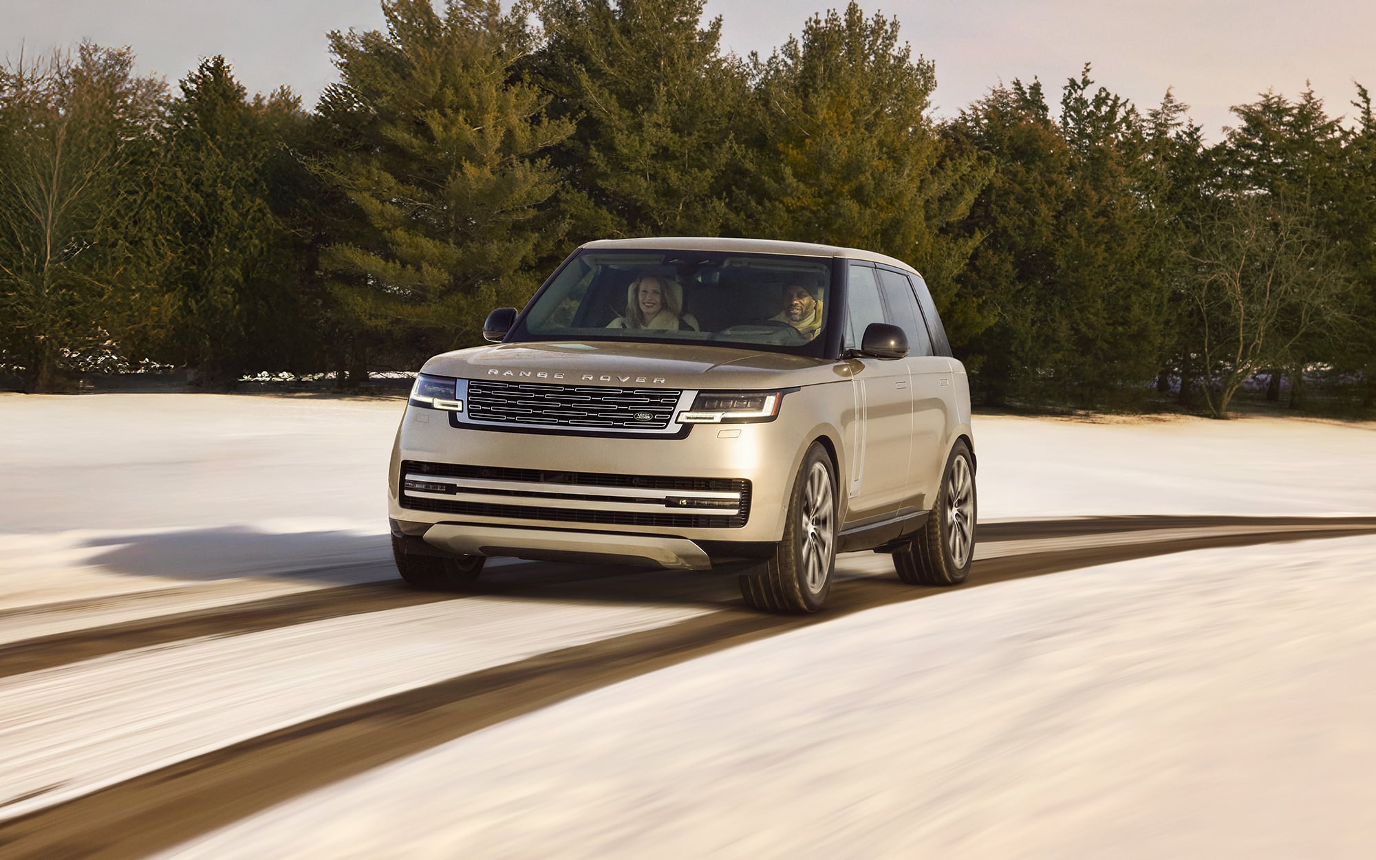 Cream-colored luxury SUV with two passengers driving on a light-colored road with evergreen trees in background