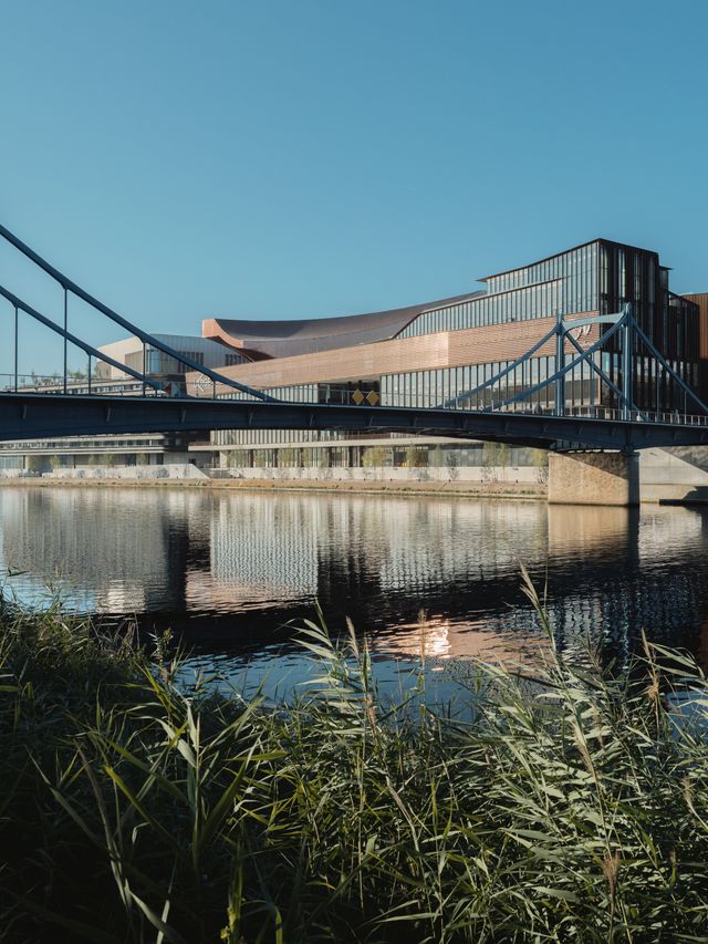 Modern waterside building with sail-like roof reflected in still water, connected by industrial footbridge