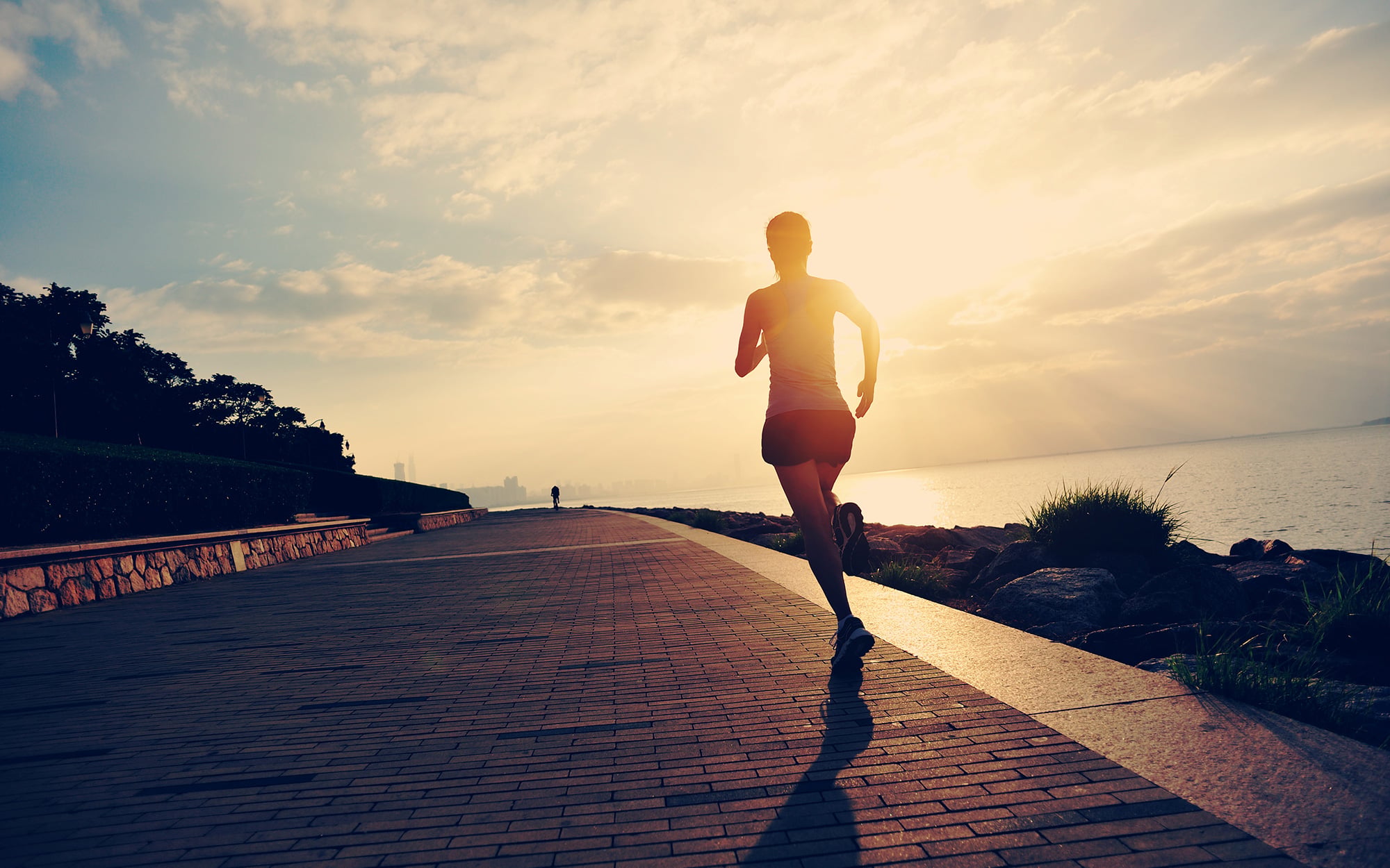 Female runner jogging on waterfront brick promenade at golden hour with ocean and sky in background