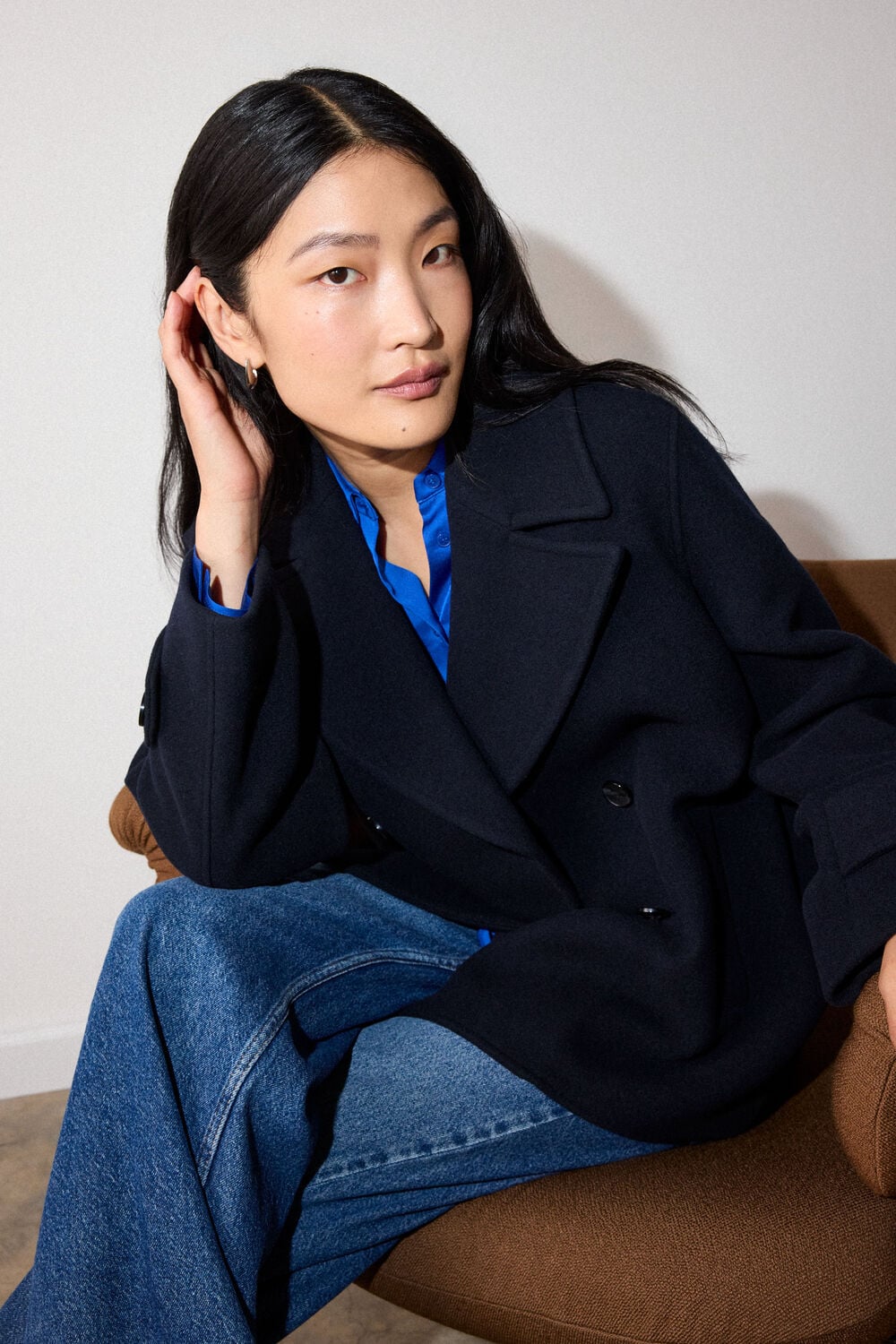 Woman with long black hair wearing navy blazer and blue shirt, sitting on brown chair with hand near ear