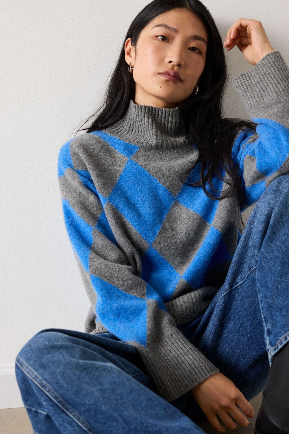Woman with long black hair wearing blue and gray argyle turtleneck sweater and jeans, adjusting hair