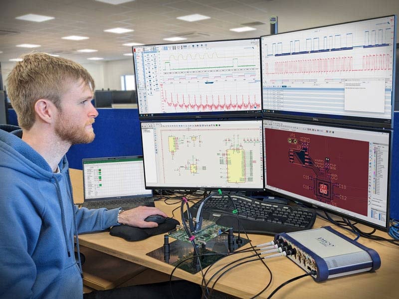 Man in blue hoodie sitting at desk with dual monitors displaying waveforms, signal analysis graphs, circuit schematics, and PCB layouts
