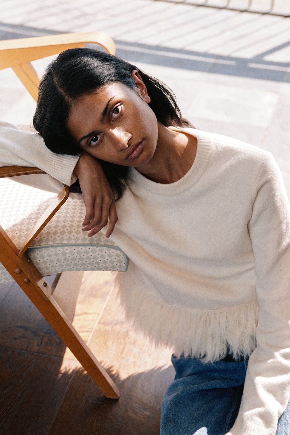 Woman with short dark hair wearing cream ribbed sweater with blue fringe trim and jeans, reclining on wooden chair