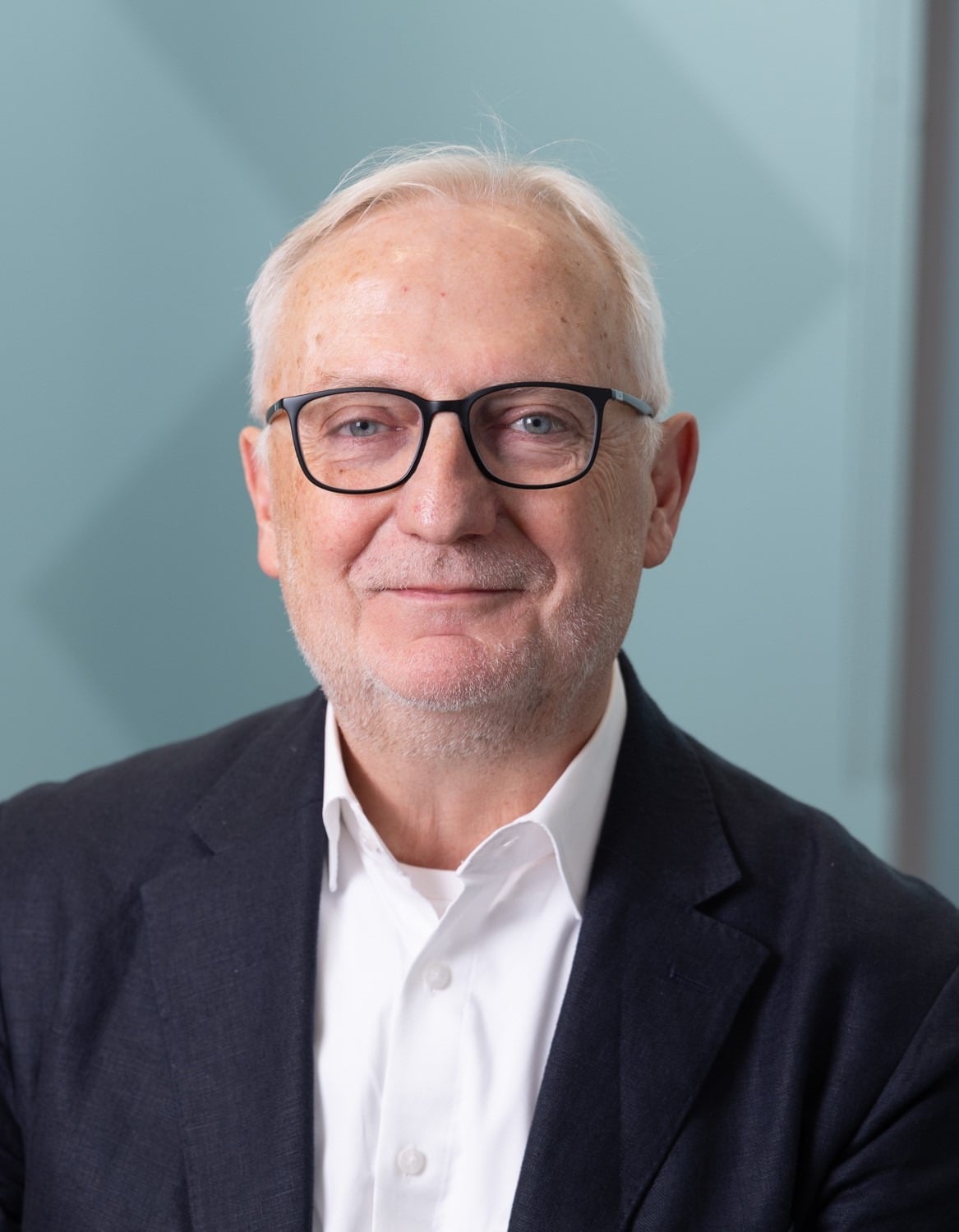 Senior man wearing glasses and dark blazer over white shirt against light blue background