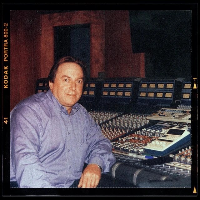 Man in purple shirt seated at professional recording studio console with multiple mixing boards and equipment