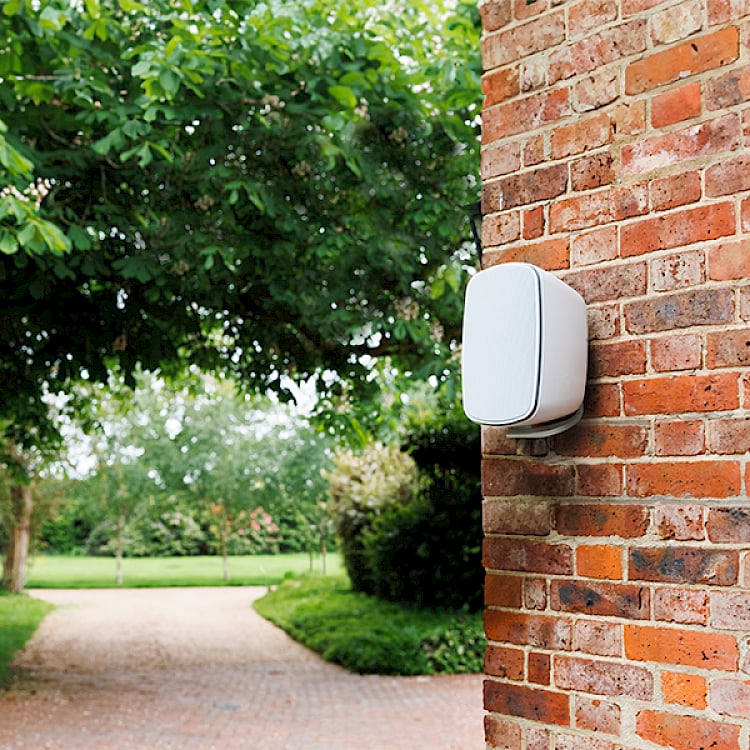 White smart home device mounted on red brick exterior wall next to green foliage