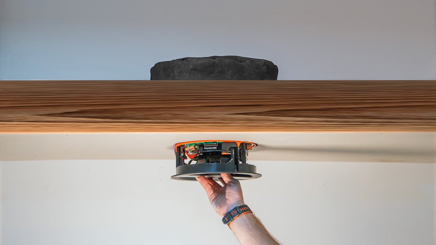 Hand holding orange and black ceiling speaker mounted on wooden beam against sky