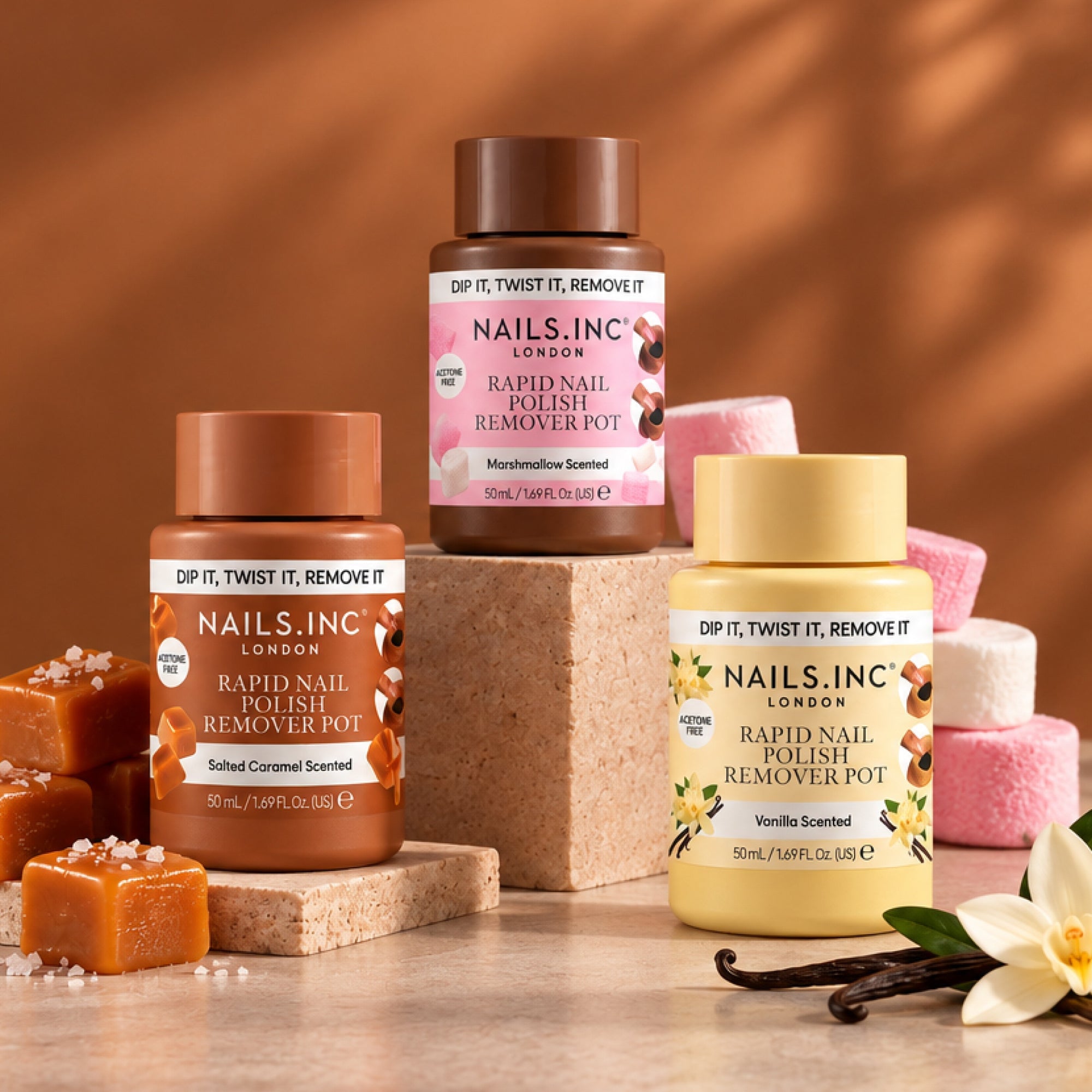 Three scented nail polish remover pots displayed on beige blocks with caramel, marshmallow, and vanilla props