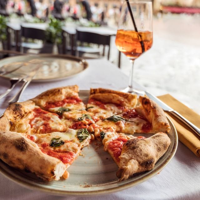 Wood-fired pizza with tomato sauce and basil served on white plate with Aperol Spritz cocktail on beachside table